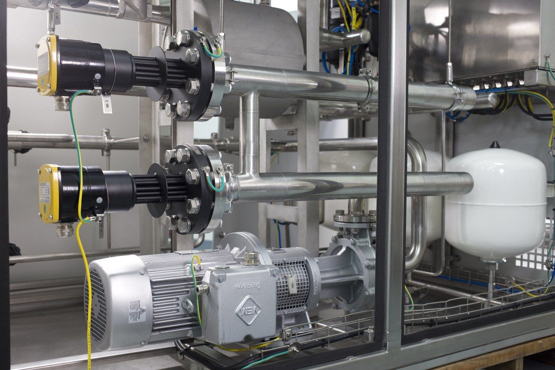 Close-up of stainless-steel process pipework, flanged connections and a pump motor inside a heat pump skid