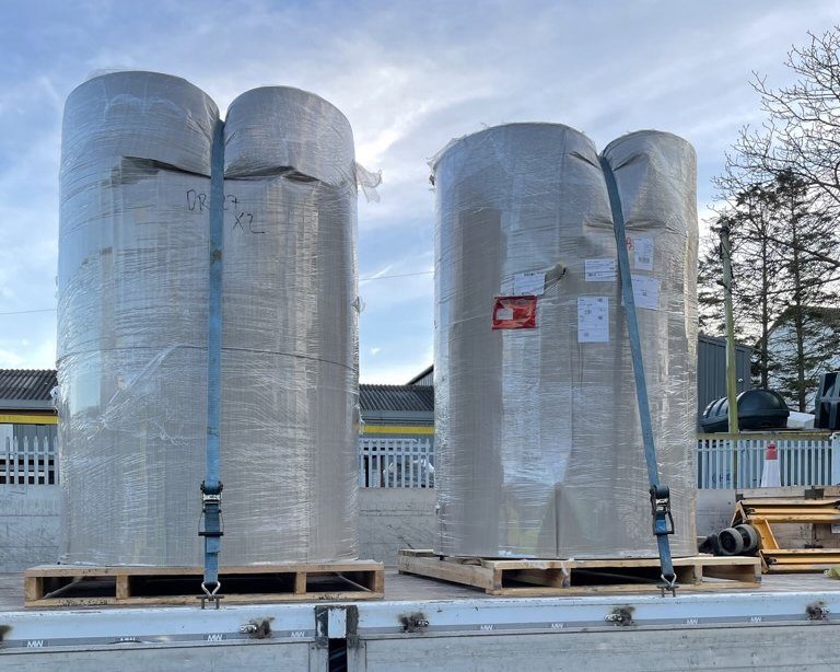Two large insulated stainless-steel hot water tanks wrapped on pallets on a flatbed truck